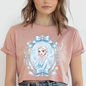 Pode incluir: Uma camiseta cropped rosa empoeirado com uma ilustra&ccedil;&atilde;o emoldurada de um personagem com cabelo branco e um vestido azul. A moldura &eacute; adornada com la&ccedil;os azuis e flocos de neve. A camisa tem mangas enroladas.