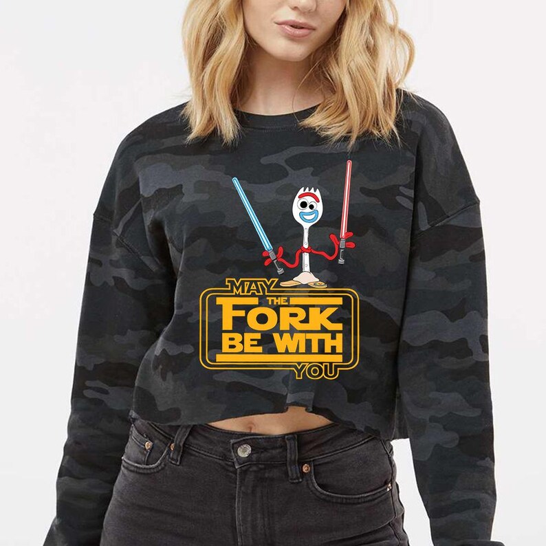 May include: A black and grey camouflage print long-sleeve sweatshirt featuring a graphic of a white spork character holding a blue and a red lightsaber. Below the character, the yellow text reads MAY THE FORK BE WITH YOU in a bold font.