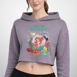 Pu&ograve; includere: Una felpa corta a maniche lunghe color malva con una stampa grafica dei personaggi de La Sirenetta, tra cui Ariel, Re Tritone, Ursula, Sebastian e Flounder. Il testo The Little Mermaid &egrave; riportato in lettere color verde acqua sopra i personaggi.