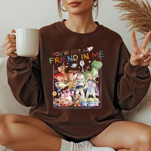 May include: A brown sweatshirt featuring a colorful graphic of characters from the movie "Toy Story." The text "YOU'VE GOT A FRIEND IN ME" is above the characters. The sweatshirt is a crew neck style.