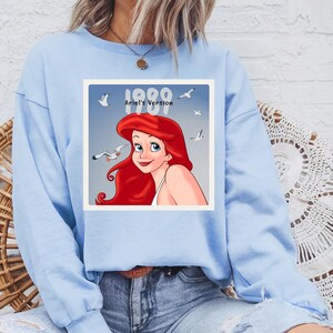May include: Light blue sweatshirt featuring a square graphic of Ariel from The Little Mermaid. The graphic includes the text "1989 Ariel's Version" with Ariel's red hair and blue eyes. White seagulls are also in the graphic.