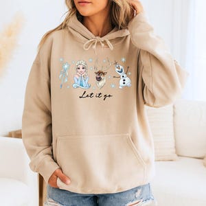 Op de afbeelding: Een beige hoodie met een afbeelding van Elsa, Sven en Olaf uit de film Frozen, met de tekst "Let it go". Het ontwerp bevat sneeuwvlokken en is gecentreerd op de voorkant. De hoodie heeft een capuchon met trekkoord en een voorzak.