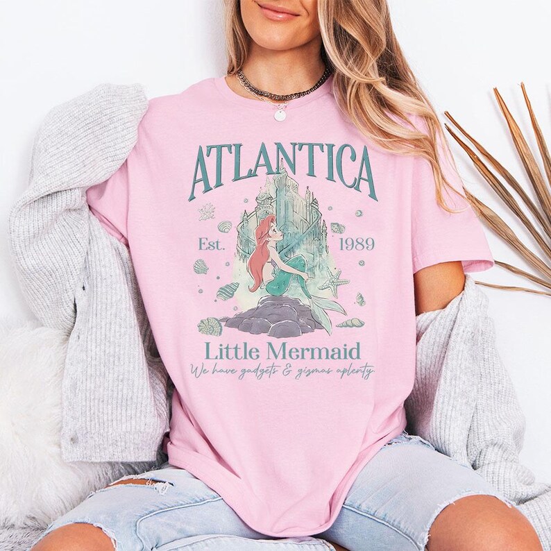 May include: Pink t-shirt featuring the text "Atlantica" above an illustration of a mermaid and castle. The shirt also includes the text "Little Mermaid" and "We have gadgets & gizmos aplenty".