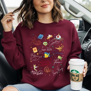 May include: A maroon crewneck sweatshirt featuring a graphic print of various fairy tale icons and character names written in cursive script, including Belle, Cinderella, Snow White, Jasmine, Rapunzel, Ariel, Fiona, Tinker Bell, and Sleeping Beauty.