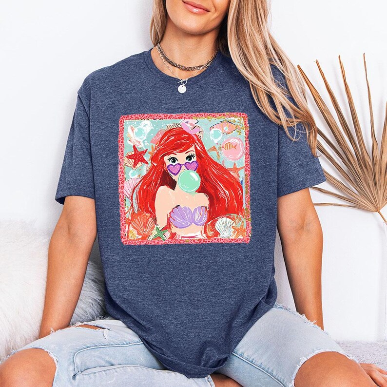 May include: Navy blue t-shirt featuring a colorful graphic of a mermaid with red hair, heart-shaped sunglasses, and a bubble. The graphic includes seashells and starfish. The shirt is a comfortable, casual style.