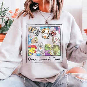 May include: A light gray sweatshirt featuring a colorful graphic of cartoon characters inside a framed rectangle. The graphic includes the text "Once Upon A Time" below the characters. The sweatshirt is made of a soft material.