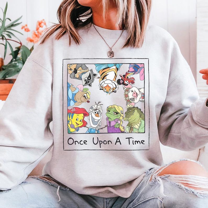 May include: A light gray sweatshirt featuring a colorful graphic of cartoon characters inside a framed rectangle. The graphic includes the text "Once Upon A Time" below the characters. The sweatshirt is made of a soft material.