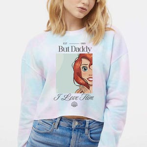 May include: A tie-dye cropped sweatshirt with a graphic of a cartoon character and the text "But Daddy I Love Him". The graphic features a red-haired character with blue eyes. The sweatshirt has a pastel color scheme.