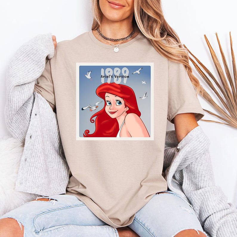 May include: Beige t-shirt featuring a square graphic of Ariel from The Little Mermaid, with red hair and a white top, surrounded by seagulls. The graphic includes the text "1989 Ariel's Version" in white.