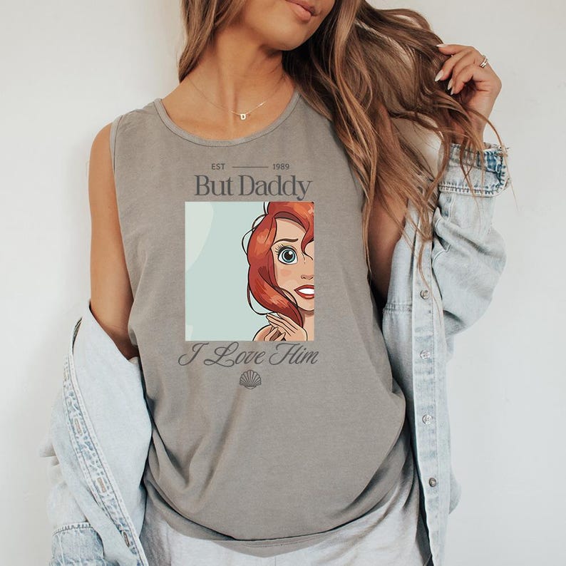 May include: Gray tank top featuring a graphic of a red-haired cartoon character with the text "But Daddy I Love Him". The tank top has a scoop neck and is made of a soft material. The graphic includes the year "1989".