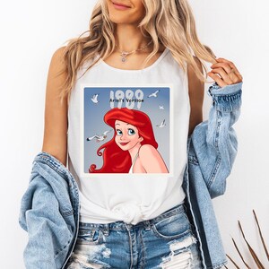 May include: White tank top featuring a graphic of Ariel from The Little Mermaid. The image includes the text "1989 Ariel's Version" with a blue background and seagulls. The tank top is worn by a person.