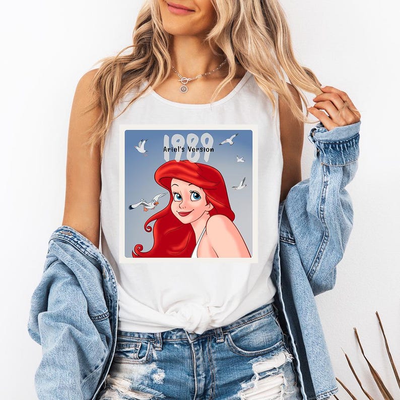 May include: White tank top featuring a graphic of Ariel from The Little Mermaid. The image includes the text "1989 Ariel's Version" with a blue background and seagulls. The tank top is worn by a person.