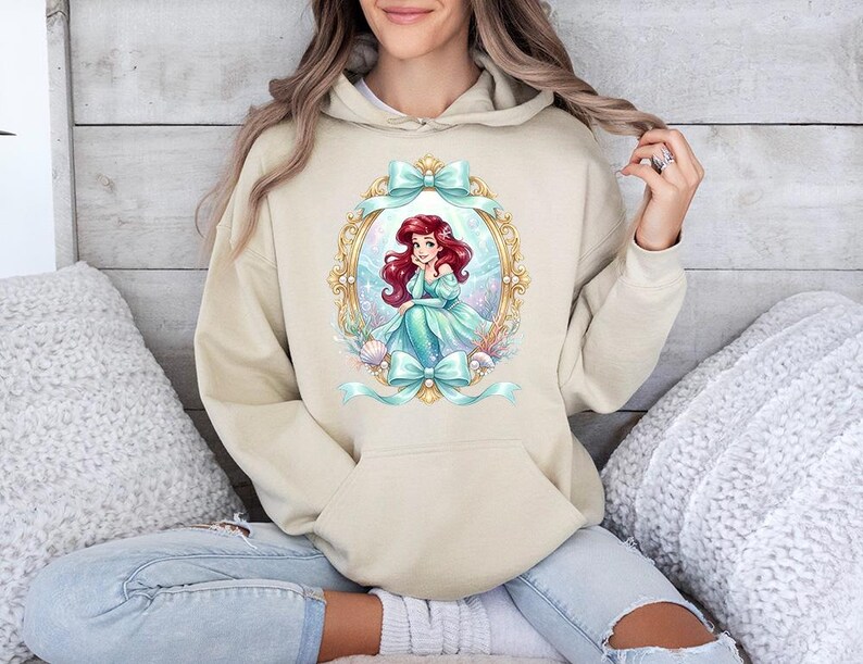 Puede incluir: Sudadera con capucha beige con una ilustraci&oacute;n de dibujos animados de una sirena en un marco ovalado dorado con lazos verde azulado. La sirena tiene el pelo rojo y lleva un vestido verde azulado. La sudadera tiene un bolsillo delantero y una capucha con cord&oacute;n.