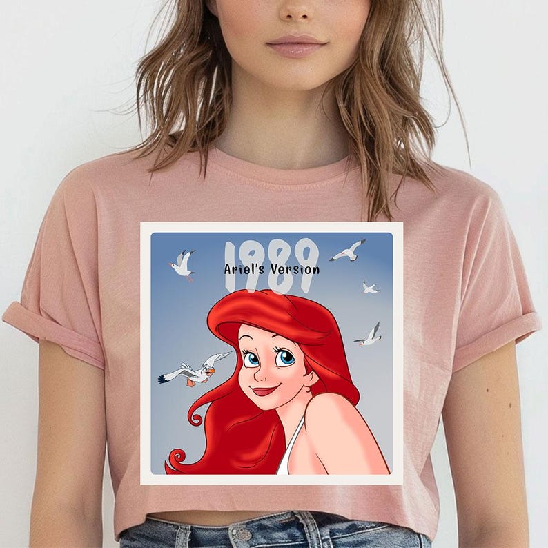 May include: A light pink t-shirt featuring a graphic of Ariel from The Little Mermaid. The graphic includes Ariel's face, red hair, and the text "1989 Ariel's Version" with seagulls in the background.