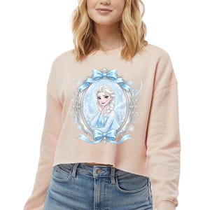 Op de afbeelding: Een lichtroze cropped sweatshirt met een ingelijste illustratie van een cartoonprinses, blauwe strikken en sneeuwvlokdetails. De prinses heeft blond haar en draagt een blauwe jurk. Het sweatshirt wordt gedragen met een blauwe spijkerbroek.