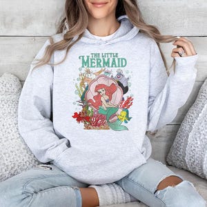 May include: A light gray hooded sweatshirt featuring a colorful graphic print of The Little Mermaid characters, including Ariel sitting in a pink seashell, King Triton, Ursula, and Flounder, with the text The Little Mermaid printed above the scene.