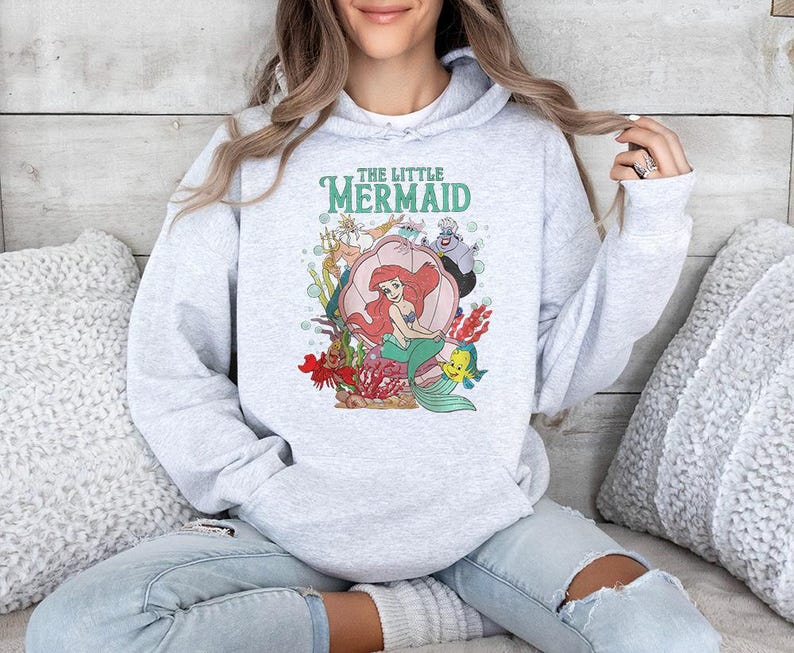 May include: A light gray hooded sweatshirt featuring a colorful graphic print of The Little Mermaid characters, including Ariel sitting in a pink seashell, King Triton, Ursula, and Flounder, with the text The Little Mermaid printed above the scene.