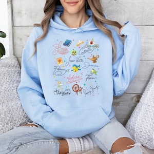 May include: A light blue hooded sweatshirt featuring a central graphic print of various character names in cursive script, including Belle, Cinderella, Snow White, Jasmine, Rapunzel, Ariel, Fiona, and Tinker Bell, alongside small icons like a book, tiara, and lamp.