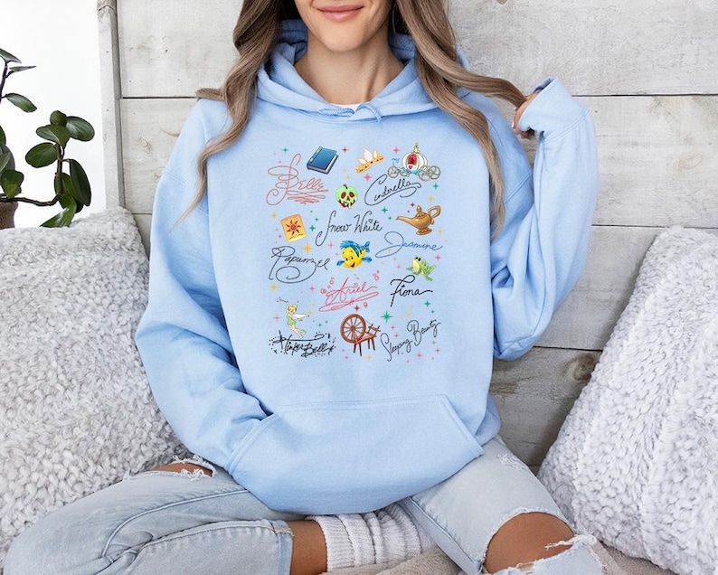 May include: A light blue hooded sweatshirt featuring a central graphic print of various character names in cursive script, including Belle, Cinderella, Snow White, Jasmine, Rapunzel, Ariel, Fiona, and Tinker Bell, alongside small icons like a book, tiara, and lamp.