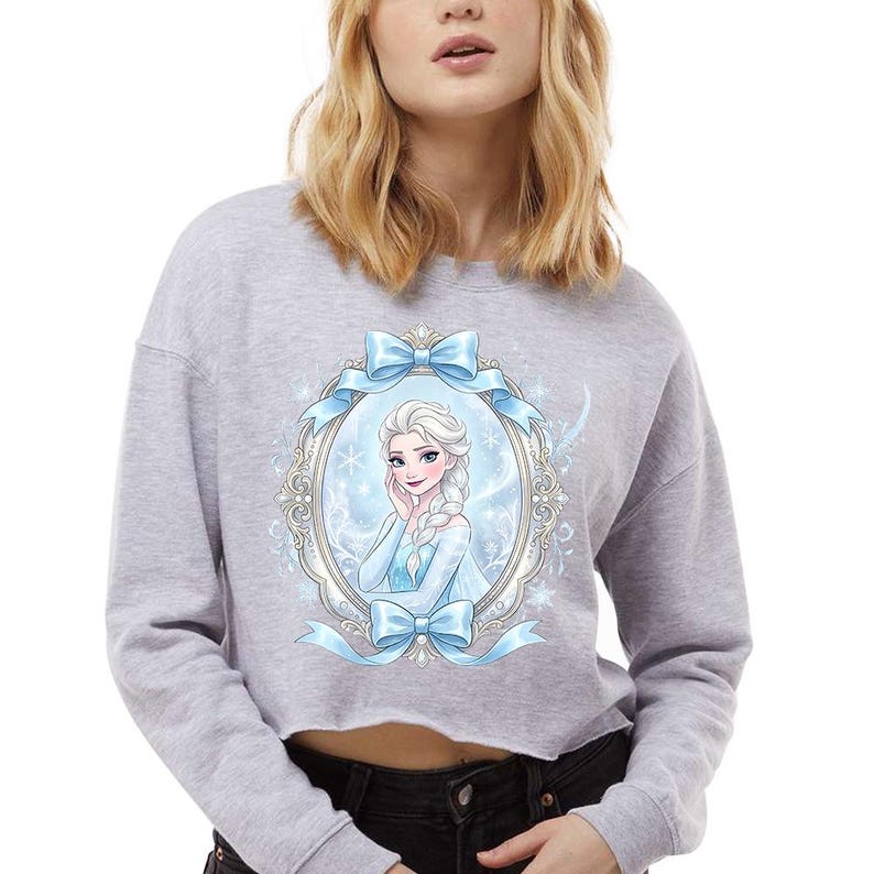 Op de afbeelding: Een lichtgrijs cropped sweatshirt met een portret van een personage met blond haar en een blauwe jurk, omlijst door een ovaal en blauwe strikken. Het ontwerp is gecentreerd op de voorkant van de sweatshirt.