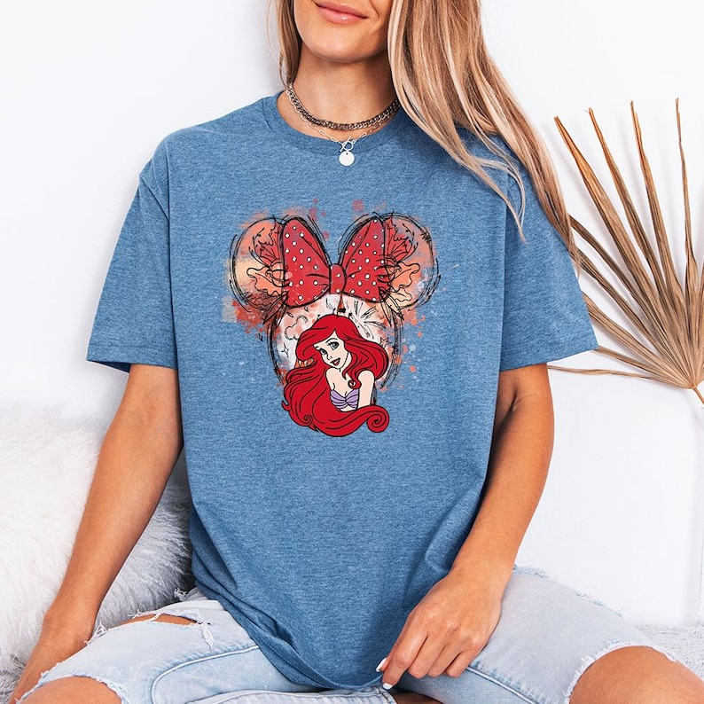 Puede incluir: Camiseta azul con un dise&ntilde;o de Ariel de La Sirenita dentro de una forma de cabeza de Mickey Mouse. El dise&ntilde;o incluye un lazo rojo con lunares y detalles de acuarela. La camiseta es de color azul liso.