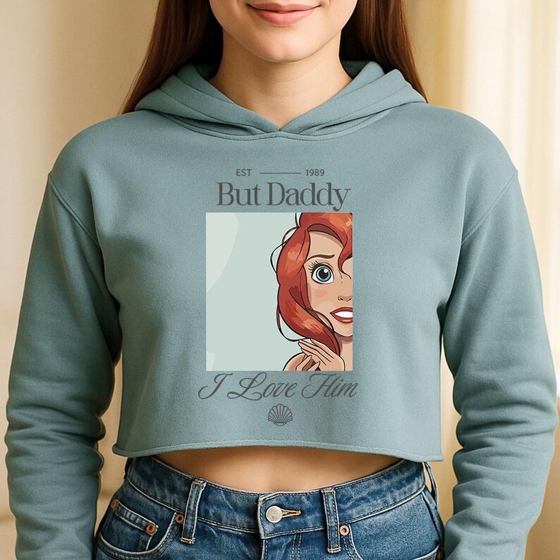 K&ouml;nnte beinhalten: Ein hellblauer, verk&uuml;rzter Kapuzenpullover mit dem Text "But Daddy" und "I Love Him". Der Hoodie zeigt eine Cartoon-Grafik einer Figur mit roten Haaren. Der Text "EST 1989" steht &uuml;ber den Worten "But Daddy".