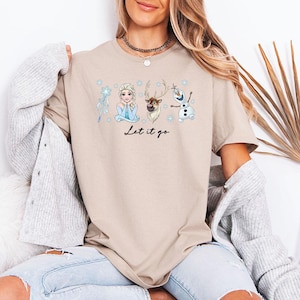 May include: Beige t-shirt with a Frozen-themed graphic including Elsa, Sven, and Olaf, plus a snowflake and wand. The words "Let it go" are written in black script. A relaxed-fit tee, suitable for everyday wear.