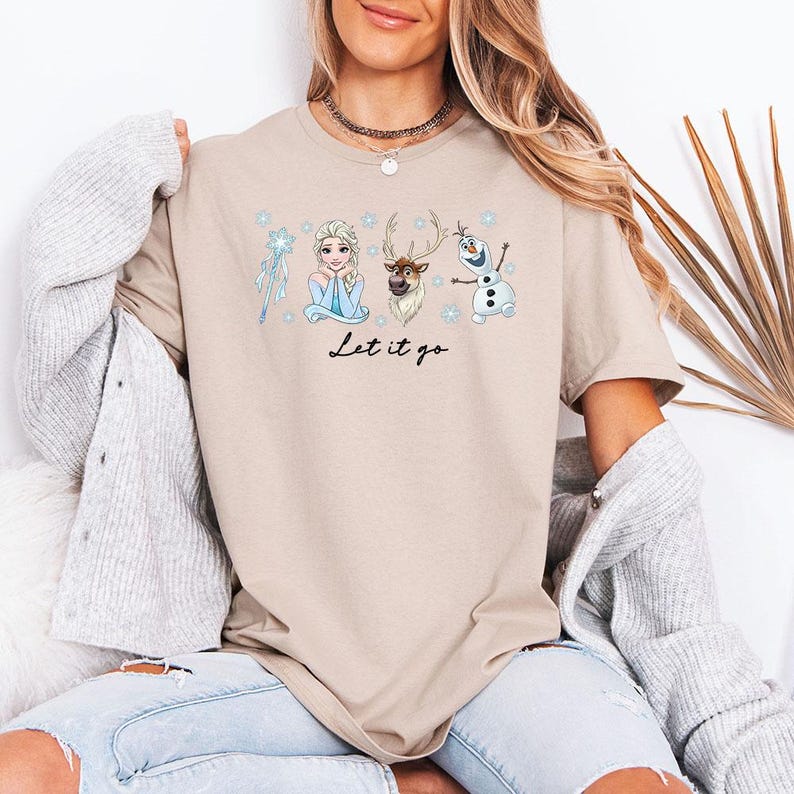May include: Beige t-shirt with a Frozen-themed graphic including Elsa, Sven, and Olaf, plus a snowflake and wand. The words "Let it go" are written in black script. A relaxed-fit tee, suitable for everyday wear.