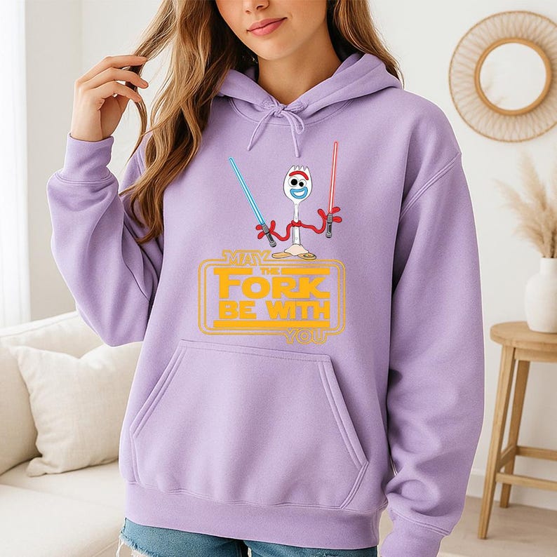 May include: A light purple hooded sweatshirt featuring a graphic of the character Forky holding a blue and a red lightsaber. Below the character, the text reads MAY THE FORK BE WITH YOU in yellow block letters. The hoodie has long sleeves and a front pocket.