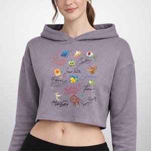 以下が含まれることがあります： モーブカラーのクロップド丈パーカー。スノーホワイト、シンデレラ、ジャスミン、ラプンツェル、アリエル、フィオナ、ティンカーベル、眠れる森の美女といったキャラクター名と、おとぎ話のアイコンがカラフルな星と共に筆記体でプリントされています。