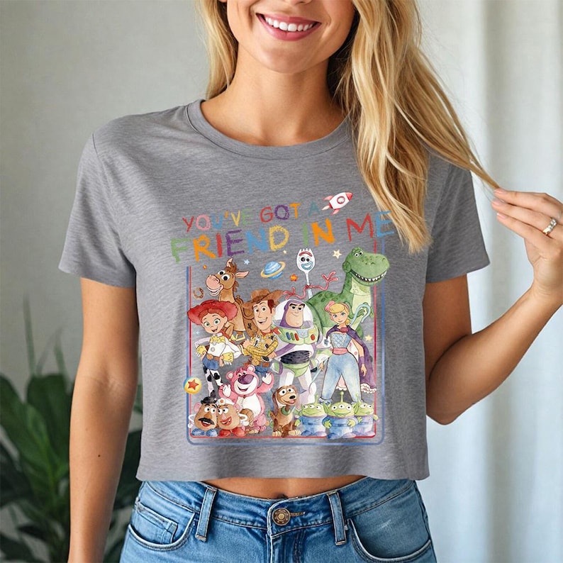 May include: Grey cropped t-shirt with a vibrant graphic of characters from the animated film "Toy Story." The design includes the text "You've Got a Friend in Me" and features characters such as Woody, Buzz Lightyear, and Rex.