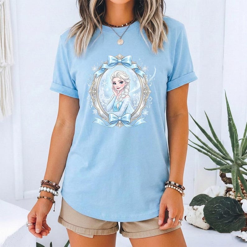 May include: Pale blue t-shirt with a framed illustration of a cartoon character with blonde hair and a blue dress. The frame is decorated with bows and snowflakes. The shirt has short sleeves.