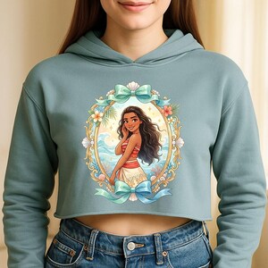K&ouml;nnte beinhalten: Ein kurz geschnittener, hellblauer Hoodie mit einer gerahmten Illustration einer Frau mit langen, dunklen Haaren, die ein rot-wei&szlig;es Outfit tr&auml;gt. Der Rahmen ist mit Blumen, Muscheln und t&uuml;rkisfarbenen Schleifen verziert. Die Frau l&auml;chelt, mit einer Strandszene im Hintergrund.