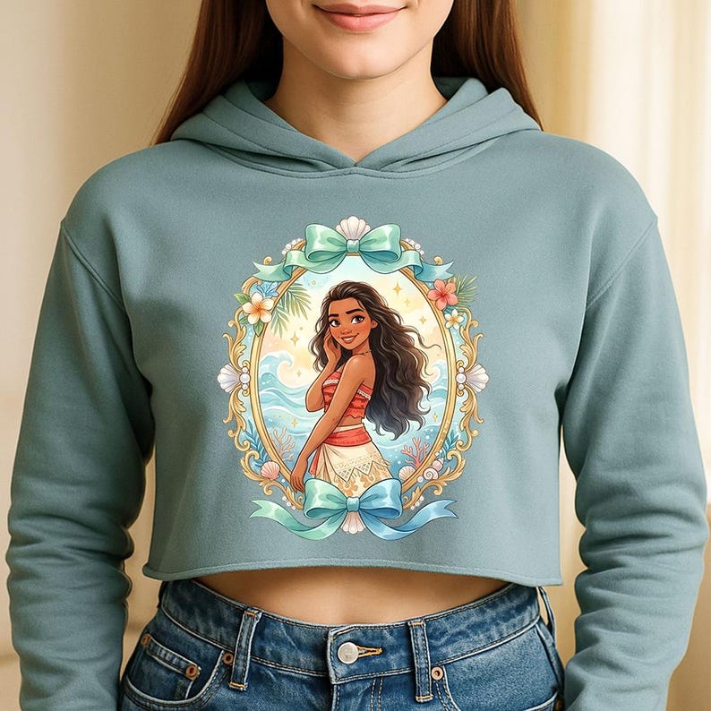 K&ouml;nnte beinhalten: Ein kurz geschnittener, hellblauer Hoodie mit einer gerahmten Illustration einer Frau mit langen, dunklen Haaren, die ein rot-wei&szlig;es Outfit tr&auml;gt. Der Rahmen ist mit Blumen, Muscheln und t&uuml;rkisfarbenen Schleifen verziert. Die Frau l&auml;chelt, mit einer Strandszene im Hintergrund.