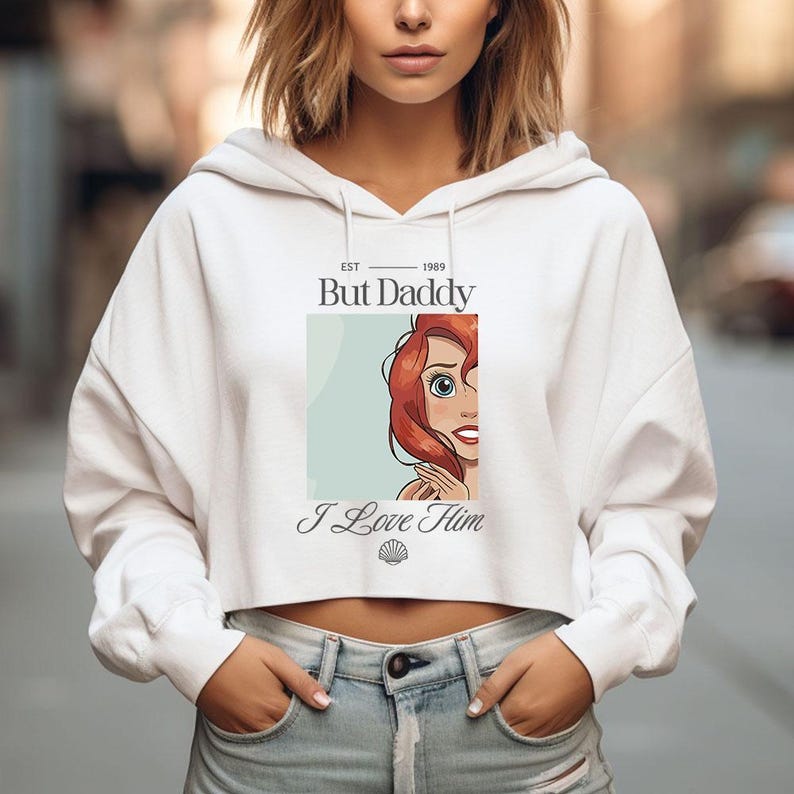 K&ouml;nnte beinhalten: Wei&szlig;er, gecroppter Hoodie mit dem Text "But Daddy" &uuml;ber einer Illustration einer Zeichentrickfigur mit roten Haaren und dem Text "I Love Him". Der Hoodie hat lange &Auml;rmel und eine Kapuze mit Kordelzug.