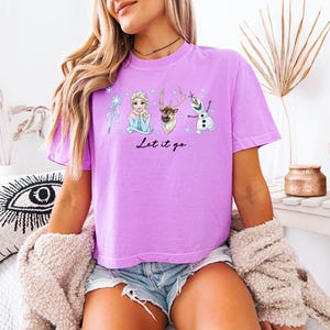May include: A lilac-colored t-shirt featuring Elsa, Sven, and Olaf from the movie Frozen. The phrase "Let it go" is written in black script below the characters. The shirt is a comfortable, casual style.