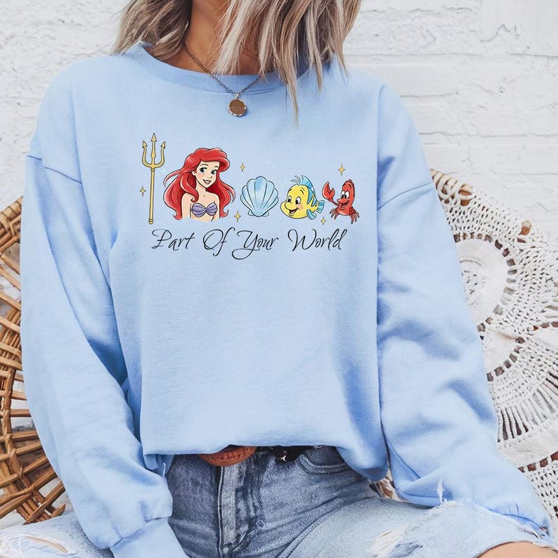 May include: Light blue sweatshirt featuring a graphic from the movie "The Little Mermaid." The design includes Ariel, Flounder, Sebastian, and a seashell, with the text "Part Of Your World."