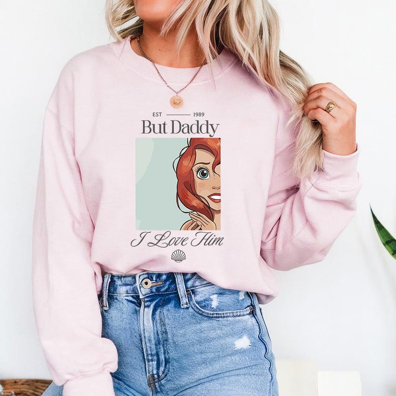 May include: A light pink sweatshirt with the text "But Daddy I Love Him" and an illustration of a cartoon character with red hair. The sweatshirt has a crew neck and long sleeves. The text "EST 1989" is above the words "But Daddy".
