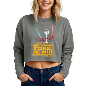 May include: A charcoal gray cropped sweatshirt featuring a graphic of a white spork character with red pipe cleaner arms holding a blue and a red lightsaber. Below the character, yellow text reads: MAY THE FORK BE WITH YOU in a rectangular frame.
