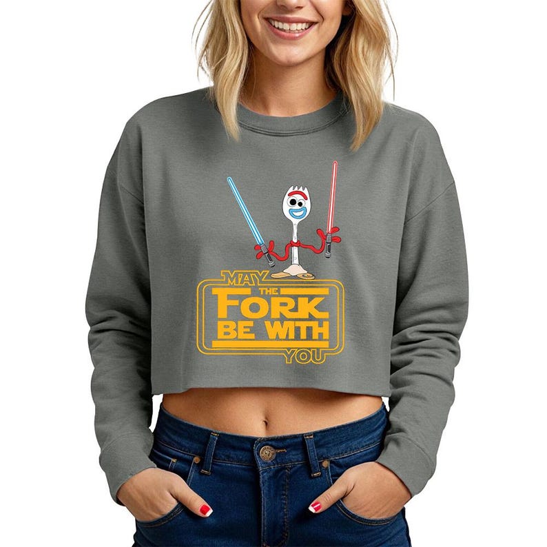 May include: A charcoal gray cropped sweatshirt featuring a graphic of a white spork character with red pipe cleaner arms holding a blue and a red lightsaber. Below the character, yellow text reads: MAY THE FORK BE WITH YOU in a rectangular frame.