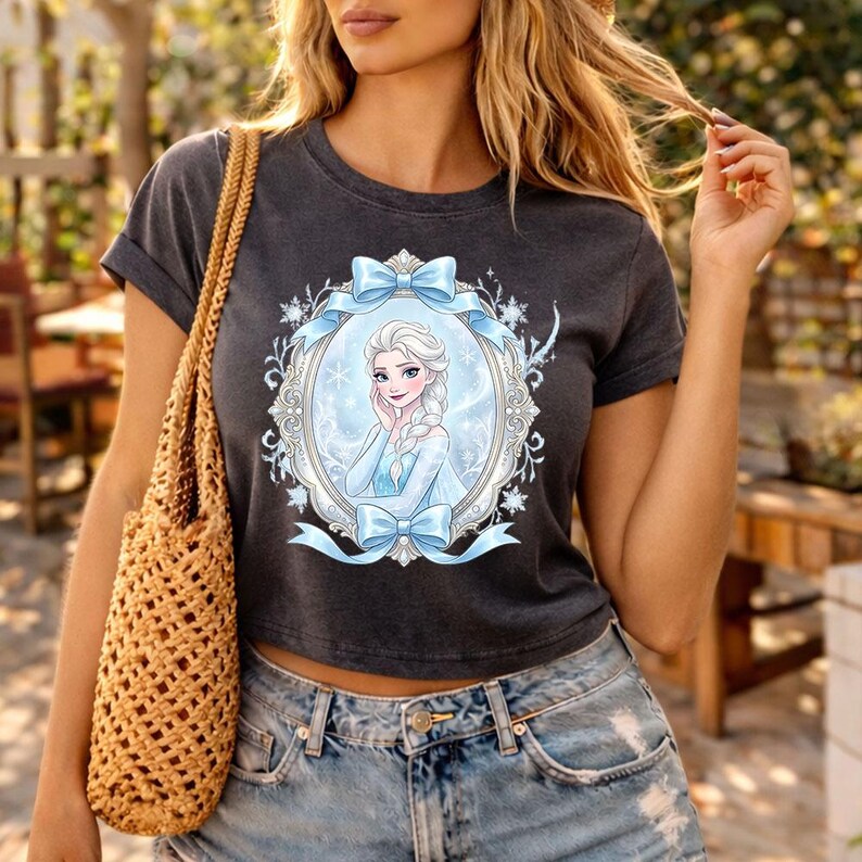Op de afbeelding: Een donkergrijze crop top met een omlijste afbeelding van een cartoonprinses in een blauwe jurk en blonde vlecht. De lijst is versierd met blauwe strikken en sneeuwvlokken. De top wordt gecombineerd met een blauwe spijkerbroek.