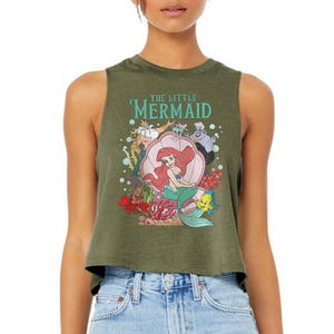 Puede incluir: Un top corto sin mangas de color verde oliva con un estampado gr&aacute;fico de los personajes de La Sirenita, incluyendo a Ariel, el Rey Trit&oacute;n, &Uacute;rsula, Sebasti&aacute;n y Flounder. El dise&ntilde;o incluye una gran concha rosa y el texto The Little Mermaid.