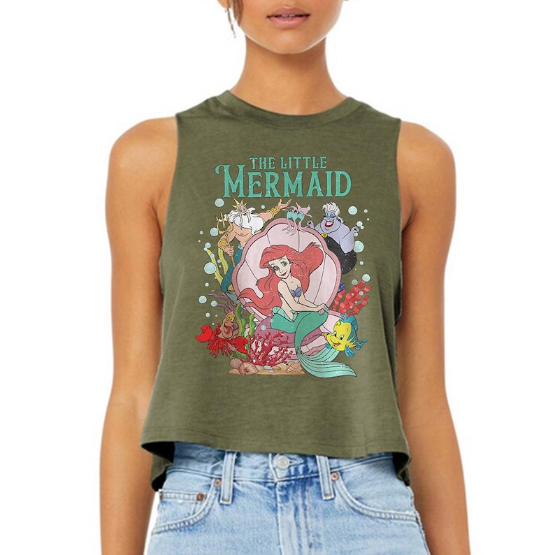 Puede incluir: Un top corto sin mangas de color verde oliva con un estampado gr&aacute;fico de los personajes de La Sirenita, incluyendo a Ariel, el Rey Trit&oacute;n, &Uacute;rsula, Sebasti&aacute;n y Flounder. El dise&ntilde;o incluye una gran concha rosa y el texto The Little Mermaid.