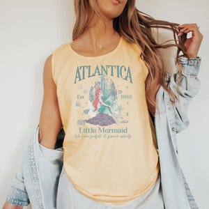 Op de afbeelding: Lichtgele tanktop met een grafische afbeelding. De afbeelding bevat het woord "Atlantica", een illustratie van een zeemeermin en de tekst "Est. 1989" en "Little Mermaid". De top is mouwloos.
