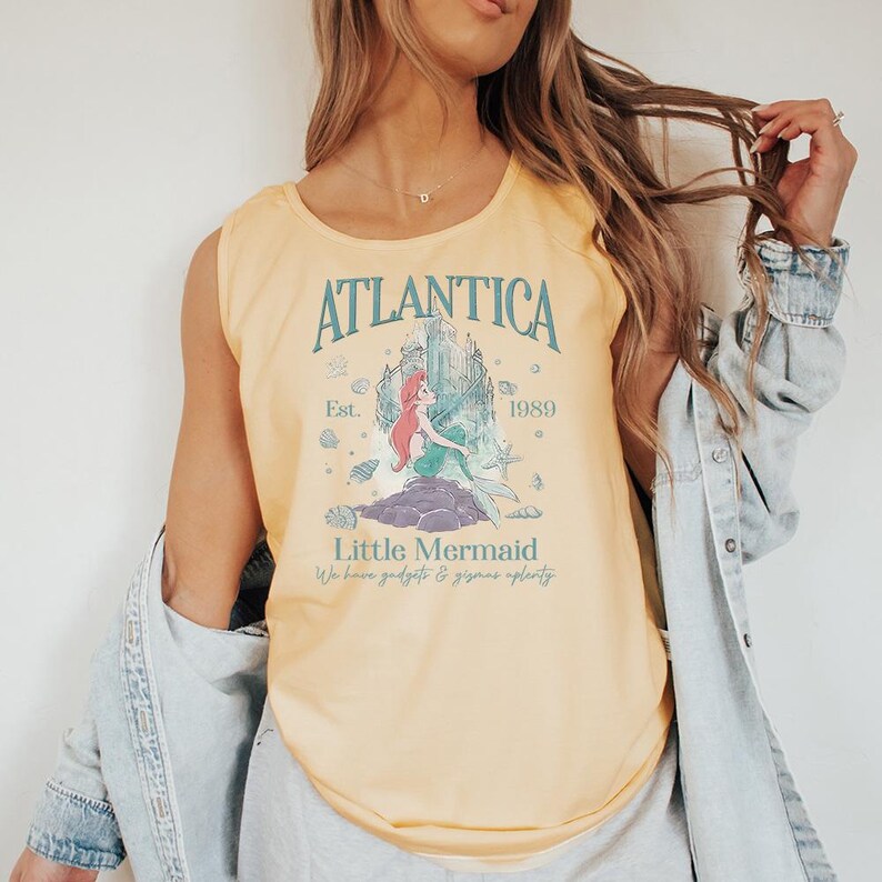 Op de afbeelding: Lichtgele tanktop met een grafische afbeelding. De afbeelding bevat het woord "Atlantica", een illustratie van een zeemeermin en de tekst "Est. 1989" en "Little Mermaid". De top is mouwloos.