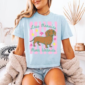 May include: A light blue t-shirt featuring a brown dachshund illustration centred on the chest. The graphic includes pink vertical stripes, colourful hand-drawn stars, and the green text "Less Meanies More Weenies" written in a cursive font.