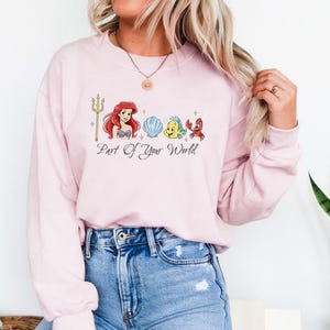May include: A light pink sweatshirt featuring a graphic of Ariel, a seashell, Flounder, and Sebastian. The text "Part Of Your World" is printed below the graphic. The sweatshirt is made of a soft material.