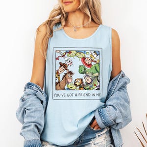 May include: Pale blue vest top with a framed illustration of Toy Story characters. The image includes Woody, Buzz Lightyear, and other characters. The text "YOU'VE GOT A FRIEND IN ME" is printed below the illustration.