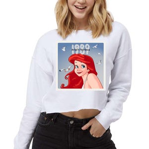Pode incluir: Camisola branca curta com um gr&aacute;fico quadrado de Ariel de A Pequena Sereia, com cabelo ruivo e fundo azul. O gr&aacute;fico inclui o texto "1989 Ariel's Version" e imagens de gaivotas.