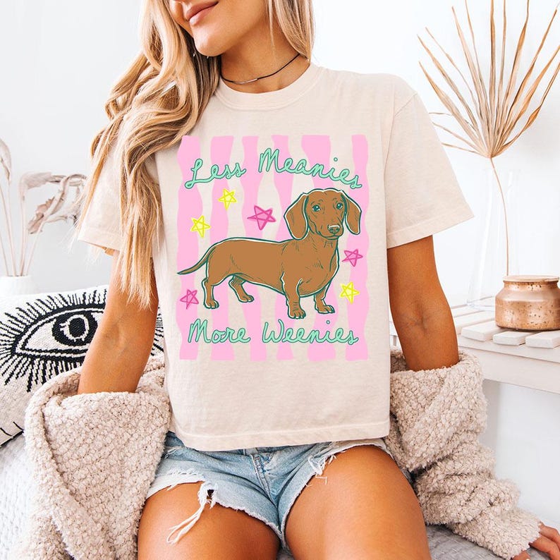 May include: A cream-coloured t-shirt featuring a brown dachshund illustration centred on a pink wavy background. The text "Less Meanies More Weenies" is printed in teal, accompanied by small yellow and pink star graphics surrounding the dog.